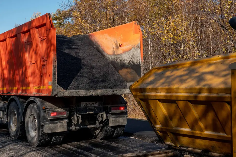 Camion-benne orange chargé, à côté d'une benne de chantier jaune, devant des arbres d'automne
